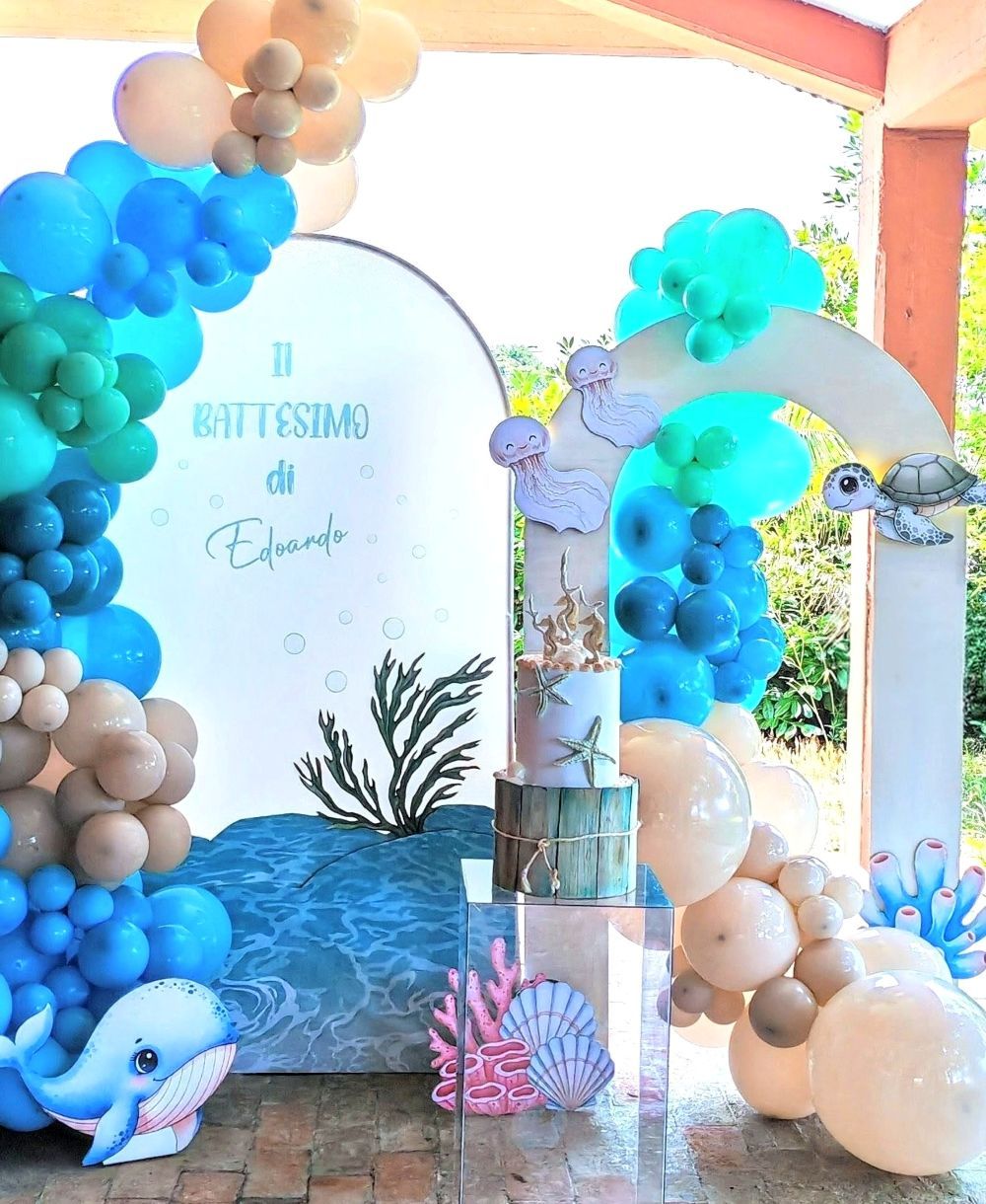 Allestimento scenografico per battesimo a tema mare realizzato da Alessia Vincenti a Roma. La scenografia comprende due strutture ad arco: quella a sinistra è decorata con un'imponente ghirlanda di palloncini organici nei toni del blu, azzurro e sabbia, ed è personalizzata con la scritta 'Il Battesimo di Edoardo'. La seconda struttura è arricchita da sagome di creature marine come una tartaruga, meduse e coralli. Al centro, su un piedistallo in acrilico trasparente, poggia una torta a due piani decorata a tema marino. Completano la composizione sagome a terra di una balena e un granchietto, il tutto su un pavimento di mattoni in un portico coperto