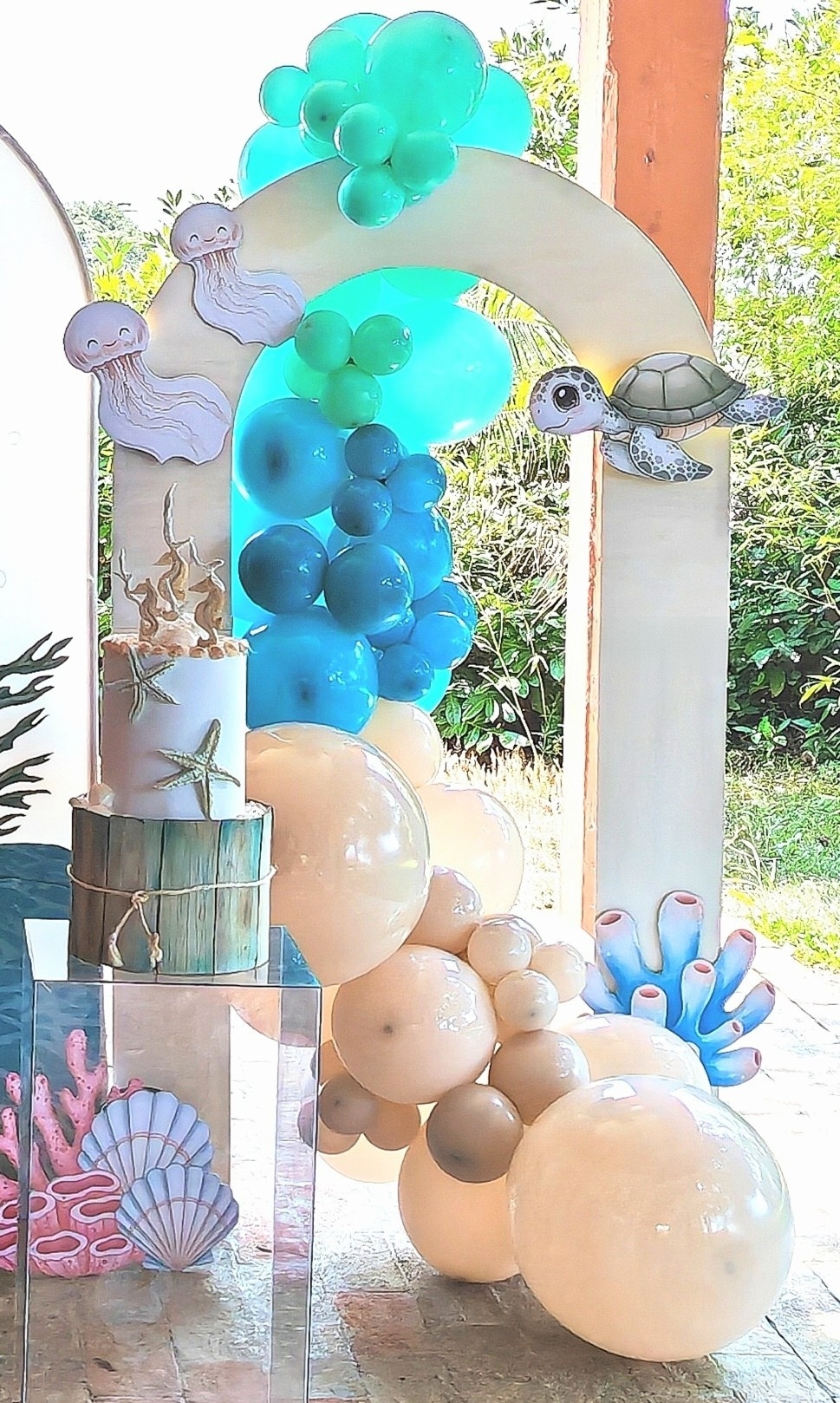 Dettaglio dell'allestimento a tema mare per battesimo, curato da Alessia Vincenti a Roma. L'immagine mostra una struttura ad arco moderna decorata con una ricca composizione di palloncini nei toni del blu, azzurro e sabbia, arricchita da sagome di creature marine, tra cui una simpatica tartaruga e meduse sorridenti. Sotto l'arco, su un piedistallo trasparente, si nota la torta a due piani, decorata con stelle marine e un topper di cavallucci marini, circondata da sagome di coralli e conchiglie colorate che completano lo scenario immersivo