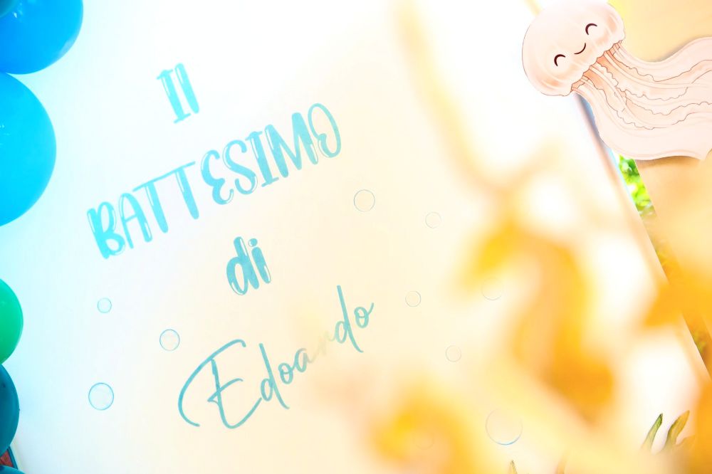 Dettaglio personalizzato di un allestimento per battesimo realizzato da Alessia Vincenti a Roma. L'immagine mostra una scritta in vinile azzurro applicata su una struttura circolare, che recita 'Il Battesimo di Edoardo'. Il design è arricchito da eleganti bolle grafiche che decorano lo sfondo e da una simpatica sagoma di medusa sorridente posizionata lateralmente, creando un'atmosfera magica e curata in ogni minimo particolare.