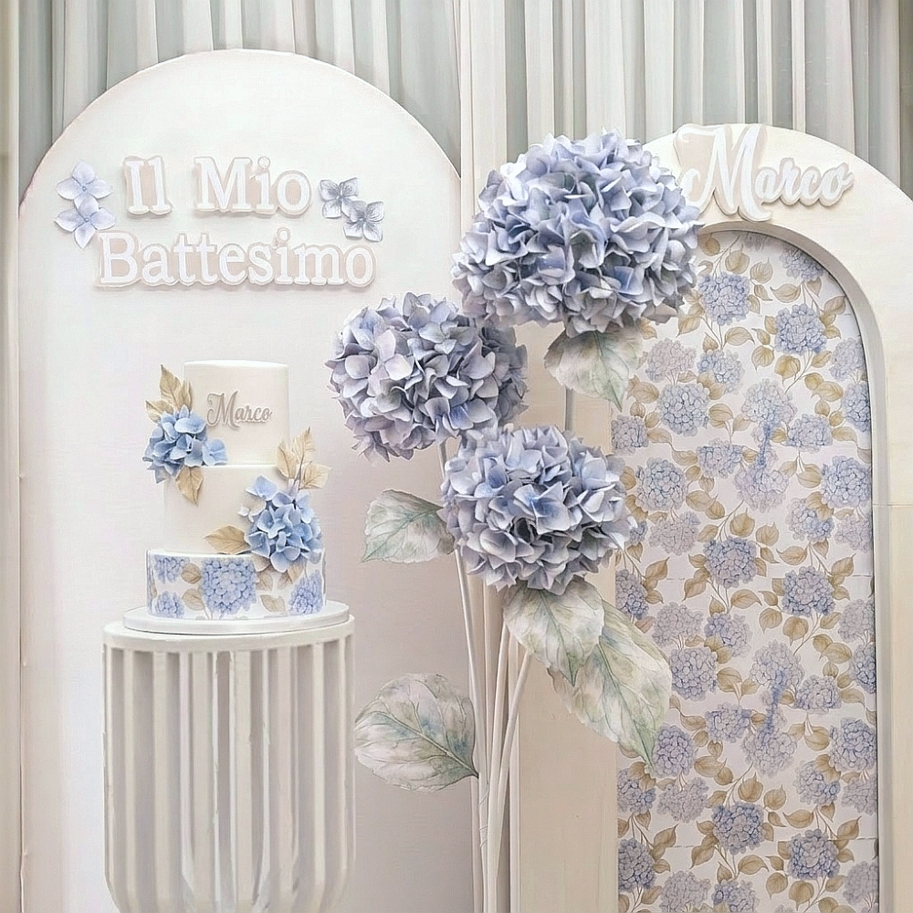 Particolare torta scenografica a tema ortensia per un battesimo con tonalità celeste polvere e beige: arco con fondale decorato, ed arco bianco con scritta in rilievo il mio battesimo, al centro fiori giganti di ortensie realizzate a mano, podio a listelli verticali. Allestimento scenografico realizzato da Alessia Vincenti Roma