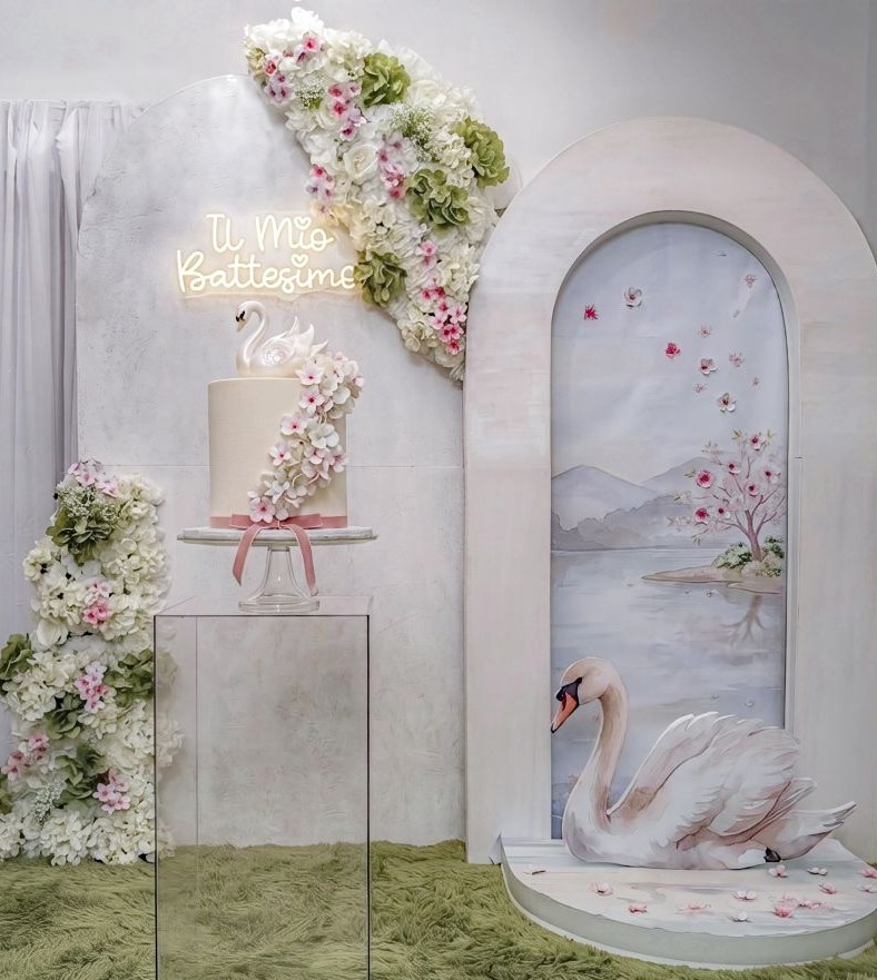 Allestimento battesimo tema cigno a Roma di Alessia Vincenti: scenografia floreale senza palloncini con cigno e sfondo realizzati tramite illustrazione digitale ad acquerello, scritta al neon Il mio Battesimo con romantico font.