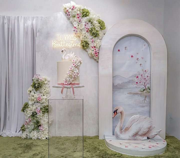 Allestimento tema battesimo con scritta al led personalizzata, composizioni fiori nei colori verde bianco e rosa, sfondo artistico di lago, sagoma cigno, torta scenografica. Allestimento, realizzato da Alessia Vincenti Roma