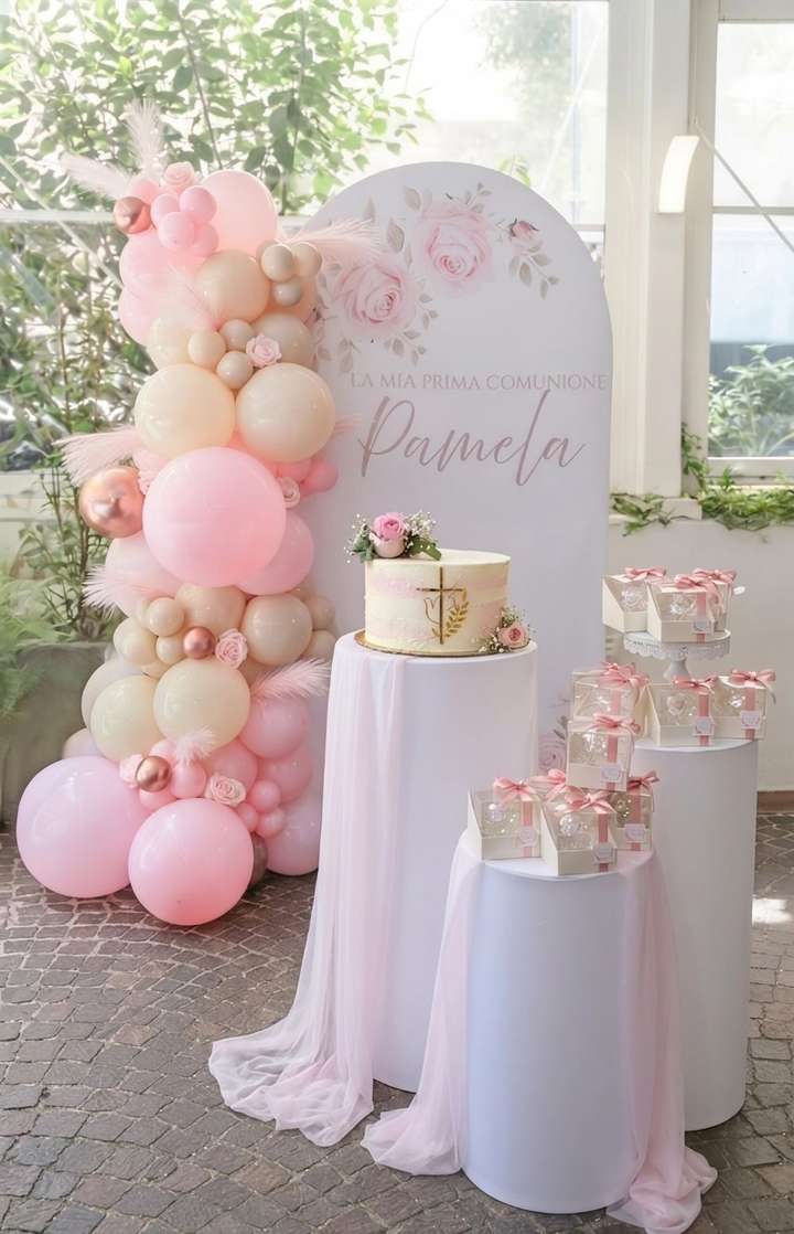 Allestimento realizzato per una comunione con grafica personalizzata, ghirlanda di palloncini nei colori beige e rosa baby, arricchita da pampas e rose, cilindri bianchi con runner rosa in chiffon, torta personalizzata e bomboniere. Allestimento realizzato da Alessia Vincenti Roma